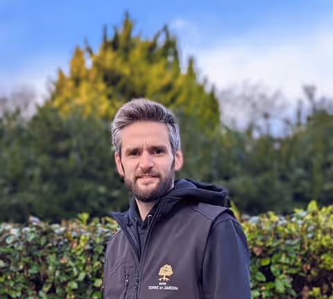 Gurvann Saintot, gérant, en gilet noir brodé du logo, devant une haie